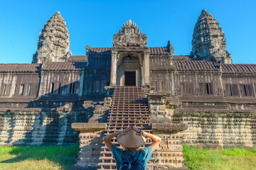 Obraz premium One tourist visiting Angkor Wat ruins at sunrise, travel destination Cambodia. Woman with traditional hat and raised arms, rear view, main facade staircase gate and towers.
