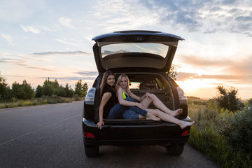 Two girls sitting in the trunk of a car. Friends relax in the summer evening and sunset behind them