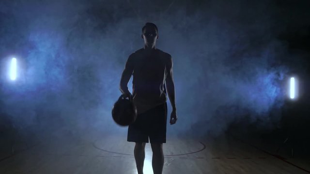 Male basketball player moving in the smoke at the camera knocking the ball about the parquet ground in slow motion Steadicam shot