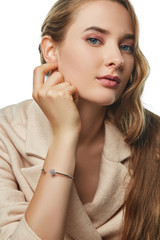 Fototapeta premium Portrait of a pretty girl with fair hair, wearing a woolen suit jacket and a bangle bracelet. The woman with fresh makeup is touching her ear while posing on white background, looking at the camera.