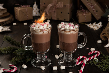Cup of hot chocolate cocoa drink with a marshmallows and sparklers and candy cane on wooden table. Two glasses of a sweet winter beverage and fir-tree branch. 