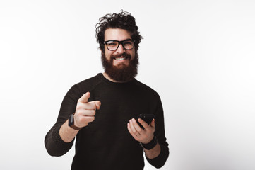 Cheerful man with beard holding smart phone and pointing at the camera