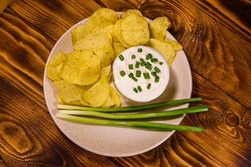 Ceramic plate with potato chips and glass bowl with sour cream on wooden table. Top view