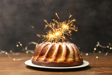 Christmas cake on white plate with sparklers on wooden table on black background with yellow lights. - Powered by Adobe