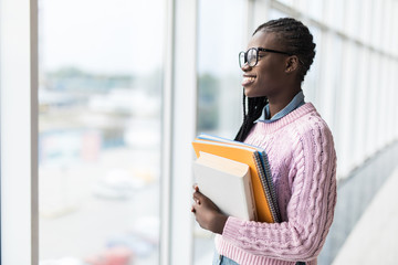 Fototapeta premium Young casual afro american student woman holding notebooks near panoramic windows in modern University