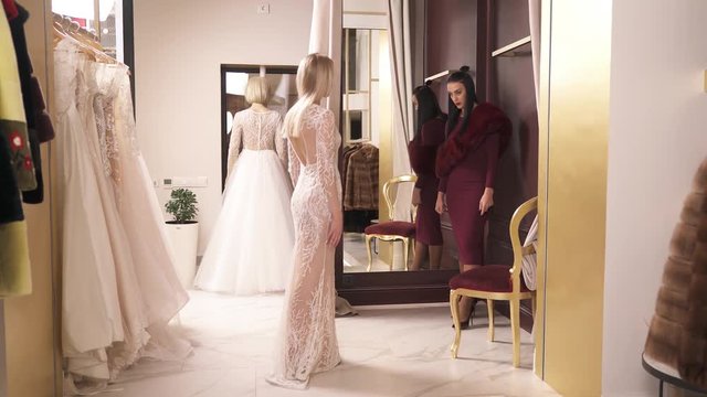 A young blonde in a long veiling fancy dress is turning around in front of the mirror and talking to the designer standing to the left. They are in a wedding dress atelier.