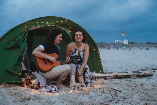 Young loving couple while camping on the seashore a summer evening at sunset. One woman plays the guitar while the other is listening to her