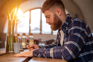 Young bearded businessman sits in cafe, home at table and writes in notebook. Man is working, studying.