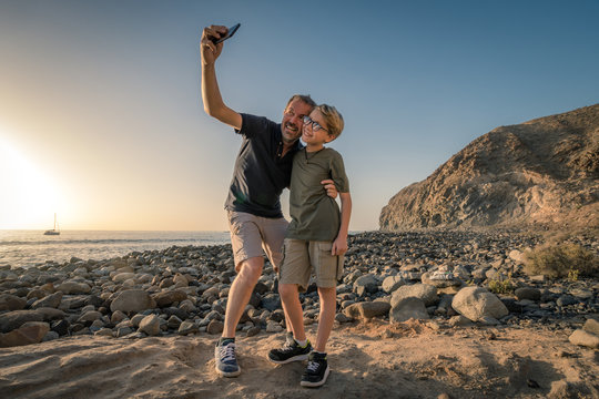 Handsome young father taking picture embracing his cute little blond son with black stare glasses on the beach, pebbles and gray stones Blue sky, calm sea sunset sailboat on the horizon Family concept