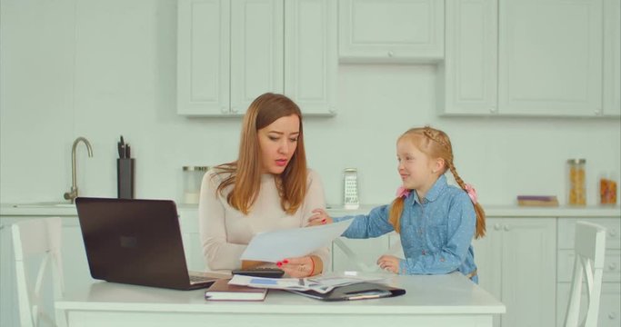 Irritated businesswoman busy working with laptop pc and paper documents ignoring her sad upset elementary age daughter in domestic kitchen. Anxious freelance mother left her child without attention.