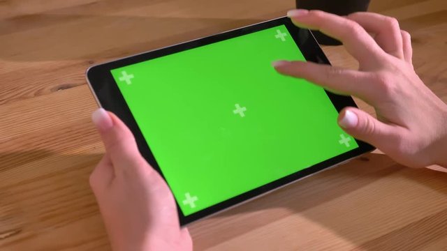 Close-up shot of horizontal tablet and hand scrolling through green screen on wooden desk background.