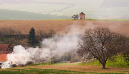 Fototapeta na wymiar Rural Pastoral Spring Landscape With Old Windmill Without Blades And Plowed Land On Background Of The Village And Thick White Smoke.European Ancient Windmill Or Defense Tower On The Brown Wavy Field