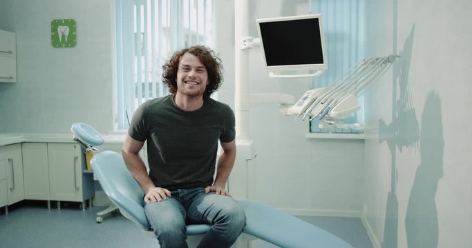 Happy and smiling patient young guy with perfect teeth sitting in the blue dentist chair and waiting for his hygiene checking , show a big like in front of the camera , in a modern white dental clinic