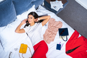 high angle view of attractive smiling girl talking by smartphone while lying on bed with stylish clothes, passport and boarding pass