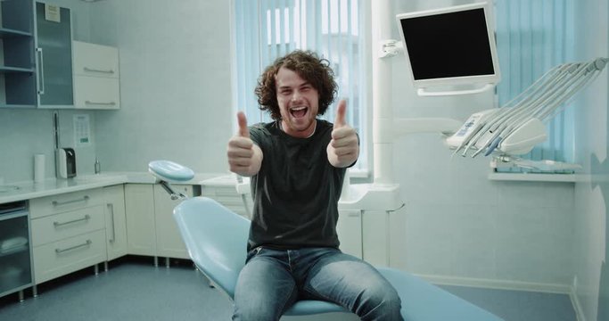 Very charismatic young patient man in a dental clinic room sitting on dentis chair after a teeth check very happy looking in front of the camera and showing a big like. 4k