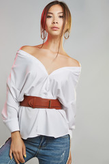 Three quarter portrait of pretty woman with tanned skin, wearing jeans and an oversized blouse with a leather belt. The lady with dark brown shin is posing on gray background, looking at the camera.