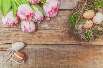 Spring greeting card. Easter eggs in nest with moss and pink fresh tulip flowers bouquet on rustic shabby wooden background. Easter concept. Flat lay top view copy space. Spring flowers tulips