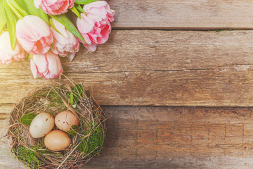 Fototapeta na wymiar Spring greeting card. Easter eggs in nest with moss and pink fresh tulip flowers bouquet on rustic shabby wooden background. Easter concept. Flat lay top view copy space. Spring flowers tulips
