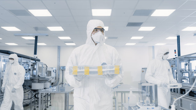 Scientist in Protective Suit Carries Case with Finished Product Through Laboratory. Facility with Modern Industrial Machinery. Product Manufacturing Process: Pharmaceutics, Biotechnology.