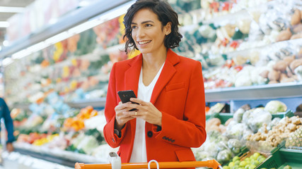 At the Supermarket: Beautiful Young Woman Uses Smartphone While Standing at the Fresh Produce Section of the Store.