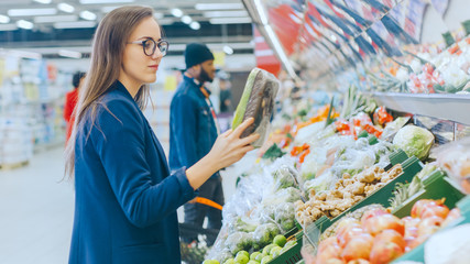 At the Supermarket: Beautiful Young Woman Stands in the Fresh Produce Section, Chooses Organic Vegetables and Fruits. Customer Shopping in the Background.