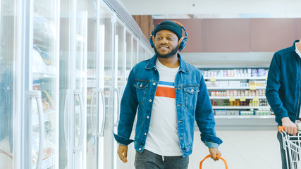 At the Supermarket: Stylish African American Guy with Headphones Walks Through Frozen Goods Section...