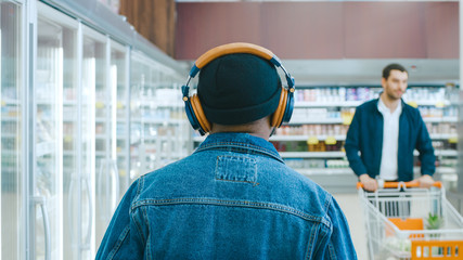 Fototapeta premium At the Supermarket: Stylish African American Guy with Headphones Walks Through Frozen Goods Section of the Store. Following Back View Shot.