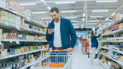 At the Supermarket: Handsome Man Uses Smartphone, Smiles while Standing at the Canned Goods Section. Has Shopping Cart with Healthy Food Items Inside. Other Customers Walking in Background.