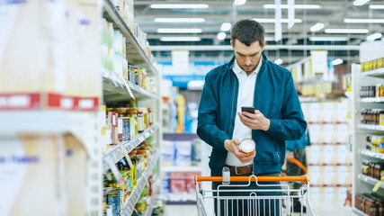 At the Supermarket: Handsome Man Uses Smartphone and Takes Picture of the Can of Goods. He's...