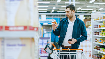 At the Supermarket: Handsome Man Uses Smartphone and Browses Through the Canned Goods Shelf. He's Standing with Shopping Cart in Canned Goods Section. - obrazy, fototapety, plakaty