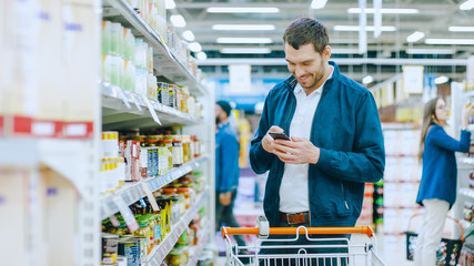 At the Supermarket: Handsome Man Uses Smartphone and Stands in Canned Goods Section. He's Looking...