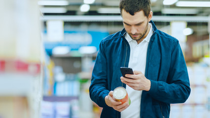 At the Supermarket: Handsome Man Uses Smartphone to Check Nutritional Value of the Canned Goods and...