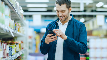 At the Supermarket: Handsome Man Uses Smartphone, Smiles while Standing at the Canned Goods...