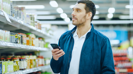 At the Supermarket: Handsome Man Uses Smartphone, Smiles while Standing at the Canned Goods Section. 