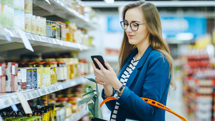 At the Supermarket: Beautiful Young Woman Uses Smartphone While Browsing through the Canned Goods Section of the Store. She Checks Her Shopping List and Holds Shopping Basket. - obrazy, fototapety, plakaty