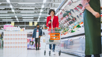 At the Supermarket: Beautiful Young Woman with Shopping Cart Uses Smartphone and Walks Through...