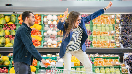 At the Supermarket: Man Pushes Shopping Cart with Woman Sitting in it, Happy Couple Has Fun Racing in a Trolley through the Fresh Produce Section of the Store.