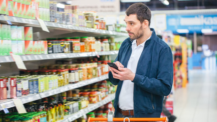 At the Supermarket: Handsome Man Uses Smartphone and Looks at Nutritional Value of the Canned...
