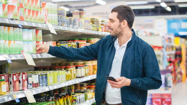 At the Supermarket: Handsome Man Uses Smartphone and Takes Tin Can. He's Standing with Shopping Cart in Canned Goods Section.