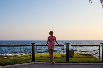 Fototapeta premium Young slender girl stands on the promenade by the sea, hot summer weather, sunset. City Side, Turkey.