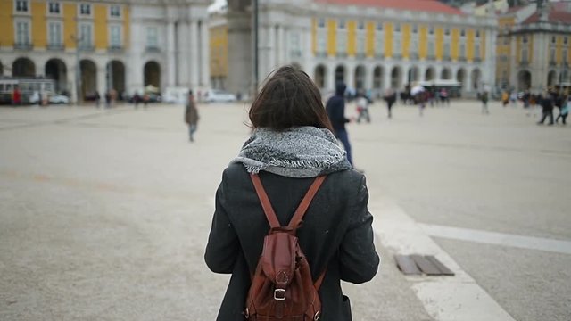 Back view of young woman with grey scarf and brown backpack walking on square