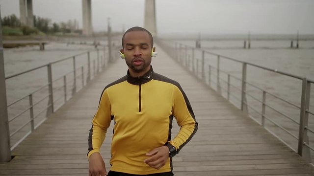 Front view of serious sporty young man running on wooden pier. Sportsman listening music with modern wireless earphones during training. Sport concept