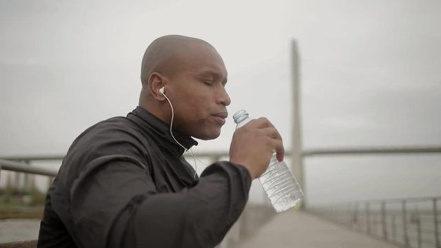 Tired sportsman drinking water while having break. Handsome young man with earphones relaxing after workout. Healthy lifestyle concept