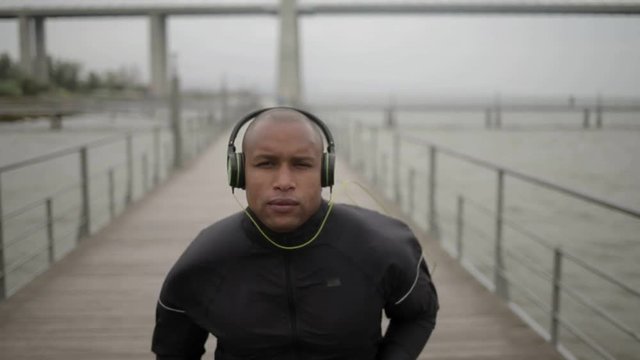 Front view of motivated bald sporty young man listening music while running on wooden pier. African american sportsman with headphones training outdoor. Training concept