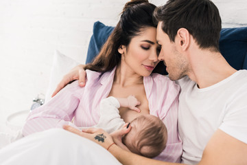 portrait of young mother breastfeeding little baby with husband near by on bed at home