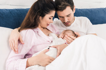 young mother breastfeeding little baby with husband near by on bed at home