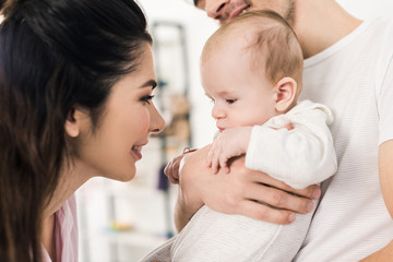 smiling mother looking at little son on fathers hands at home