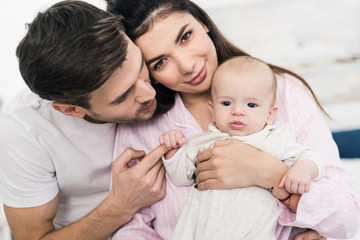 portrait of man smiling hugging wife with little son on hands at home