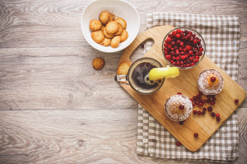 On a light wooden tabletop on a linen napkin napkin, there is a cutting board with two muffins, a broken chocolate bar and bright red berries in a small tree, next to a bowl with cookies.