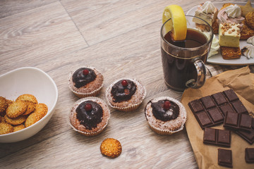 On a light wooden tabletop, four chocolate-covered cupcakes, decorated with red berries, black coffee, decorated with a slice of lemon and biscuits.